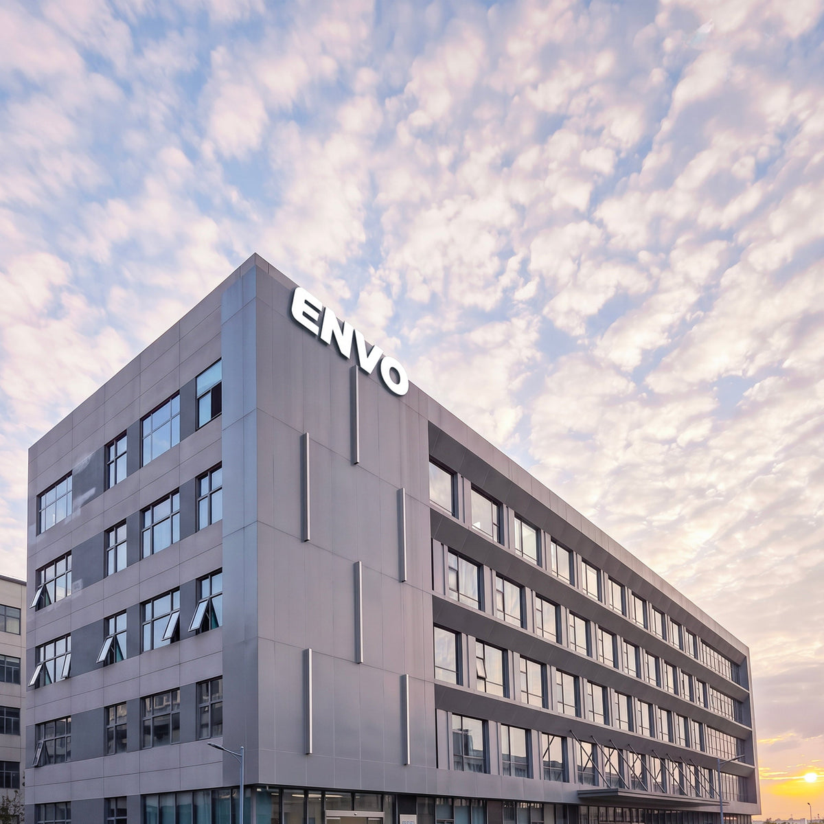 A modern multi-story office building with the brand name ENVO prominently displayed on the facade, set against a dramatic sky filled with textured clouds at sunset.