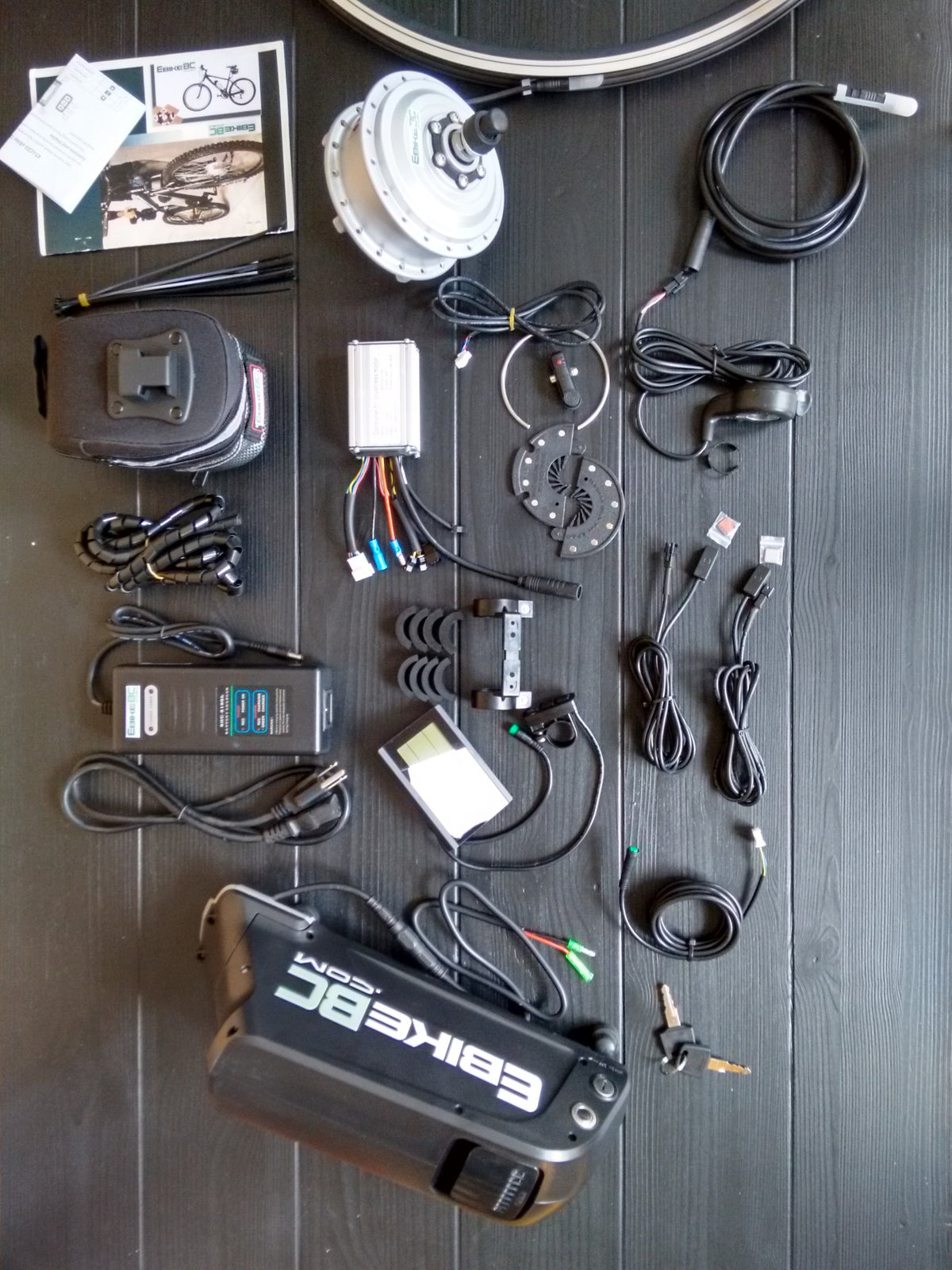 A laid-out electric bicycle battery and accessories on a dark wood surface, including a large black battery pack labeled EBIKE, a motor, cables, connectors, a charger, keys, and a small instruction booklet.